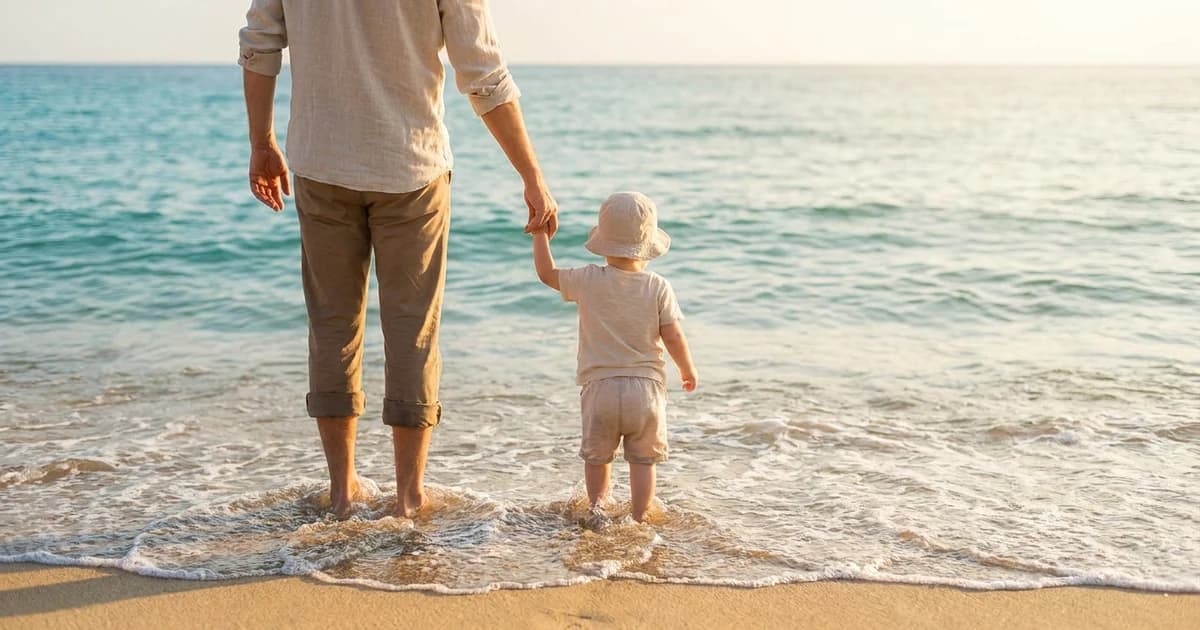 Parent holding a toddler's hand at the edge of the ocean in soft daylight as small waves reach the shore