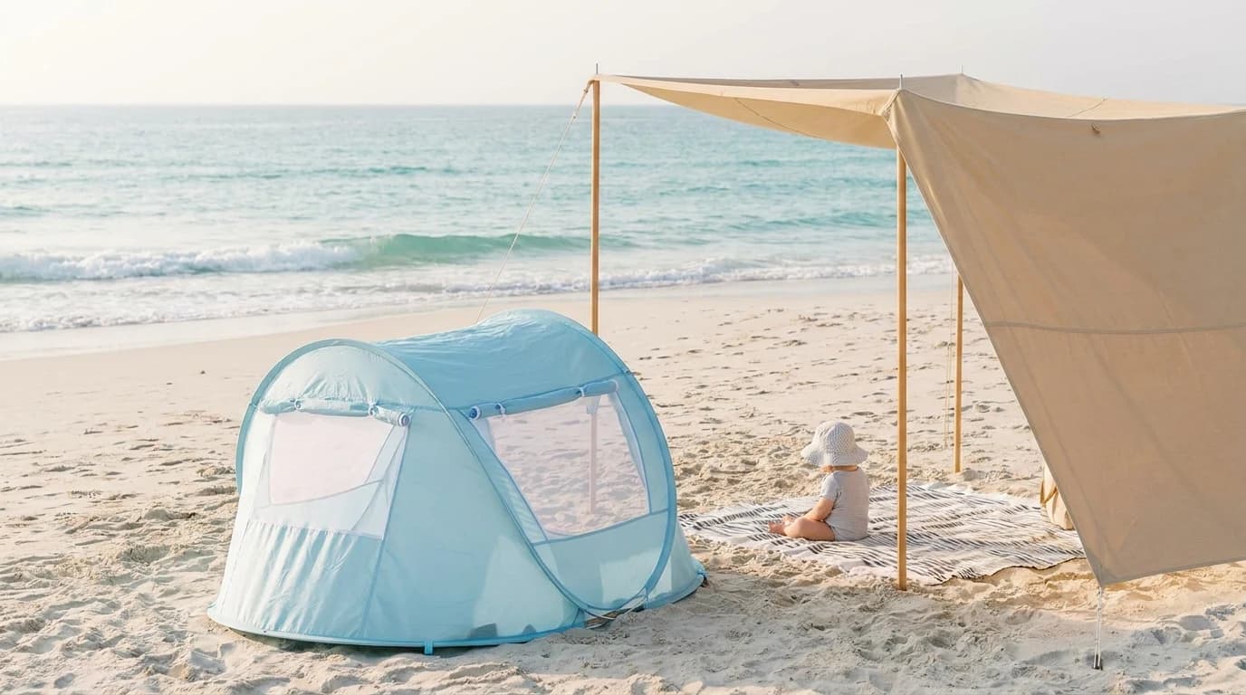 A small pop-up baby beach tent on pale sand beside a family shade canopy, with a baby in a sun hat sitting in the shade