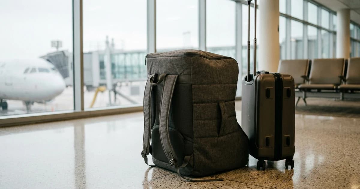 A padded grey car seat travel bag with backpack straps standing beside a rolling carry-on suitcase on the floor of an airport departure gate, an aircraft visible through the windows.