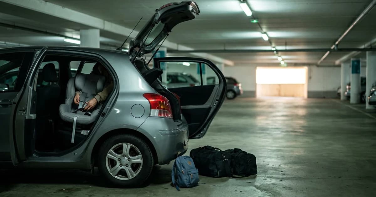 A child's car seat strapped into the back of a small European hatchback in an airport parking garage, rental luggage beside the open door in flat overhead light.