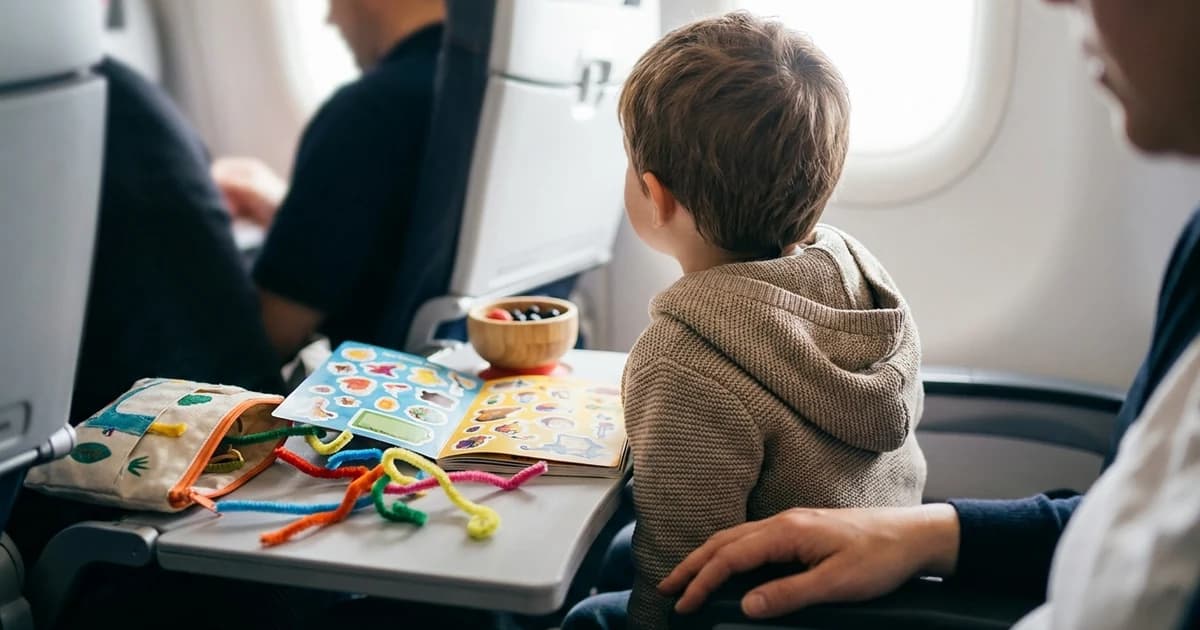 Young child playing with a reusable sticker book on an airplane tray table during a flight, no screens in sight