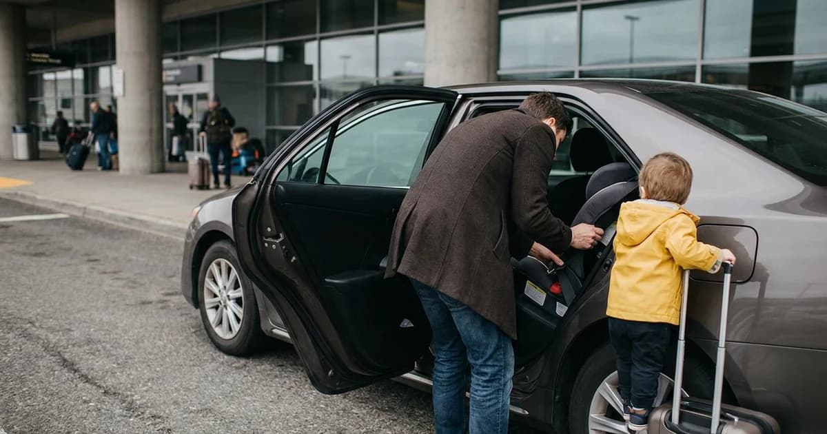 A parent buckling a travel car seat into the back seat of a grey sedan at an airport arrivals curb while a toddler in a yellow rain jacket waits beside a carry-on suitcase.