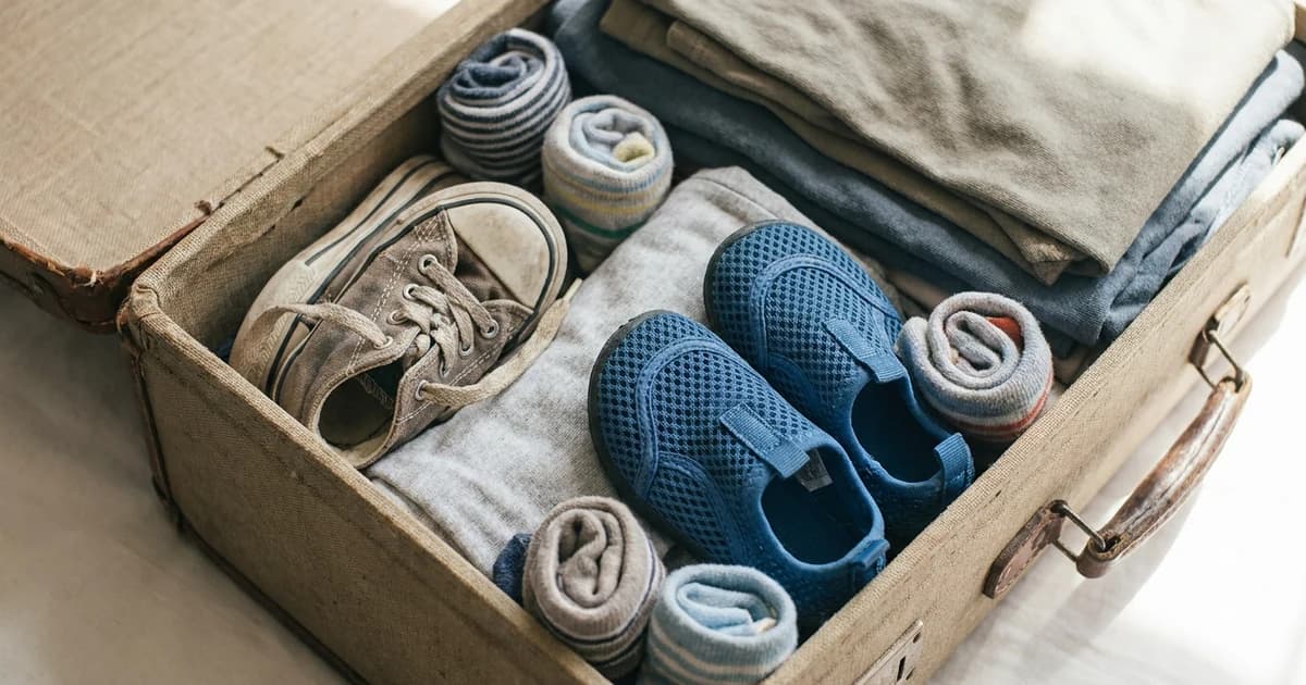 Two pairs of kids' shoes packed in an open suitcase corner — a worn closed sneaker and small closed-toe water shoes — with rolled socks tucked beside them, soft natural window light