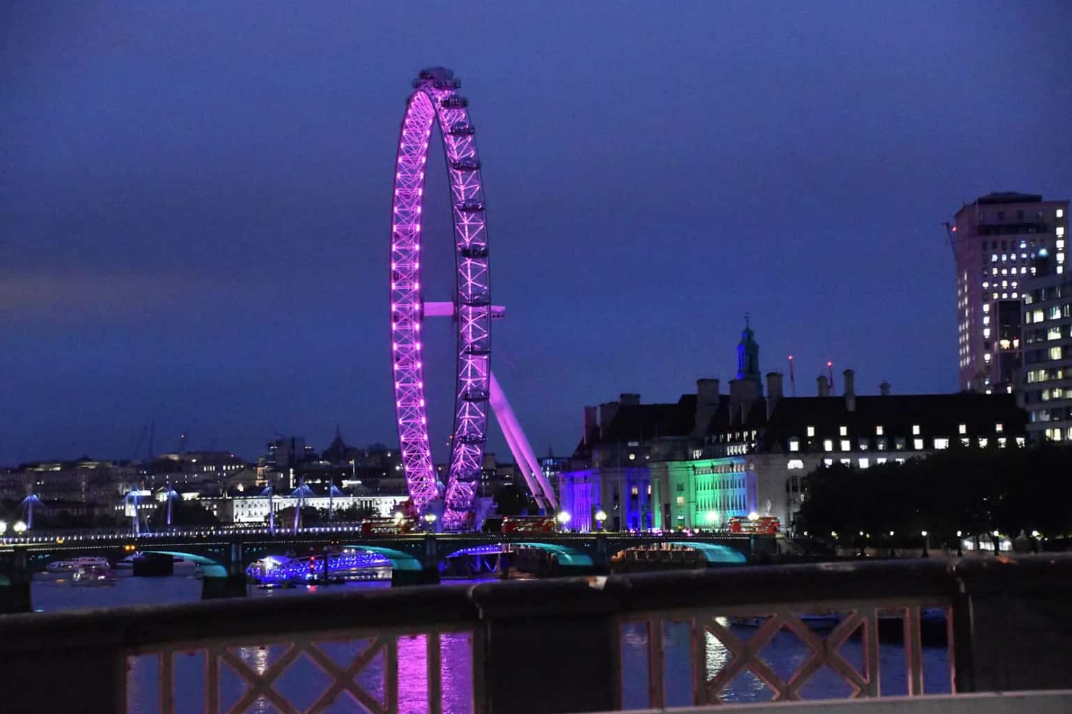 The London Eye lit up in pink and purple at night, reflected beside a bridge over the Thames.