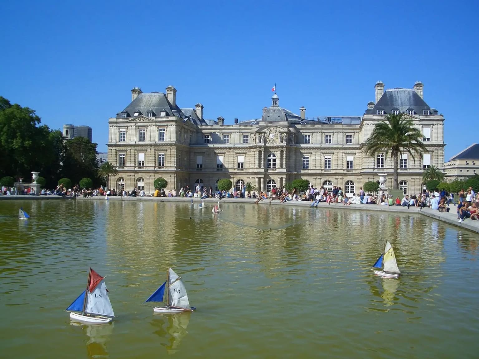 Small wooden toy sailboats with coloured sails on the Grand Bassin, with the Luxembourg Palace and visitors behind.