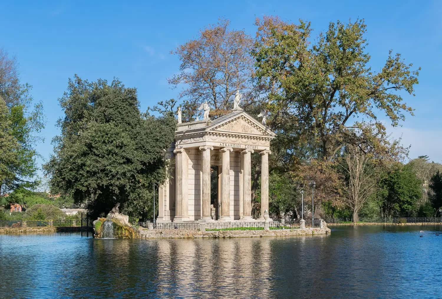 The small columned Temple of Aesculapius on an island in the Villa Borghese lake, framed by tall trees.