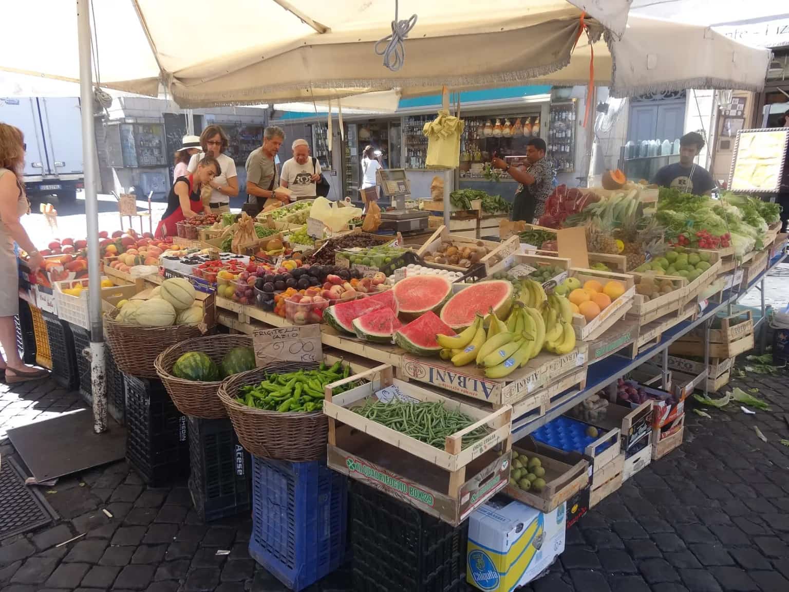 A Campo de' Fiori market stall piled with watermelons, bananas and fruit under a white umbrella.