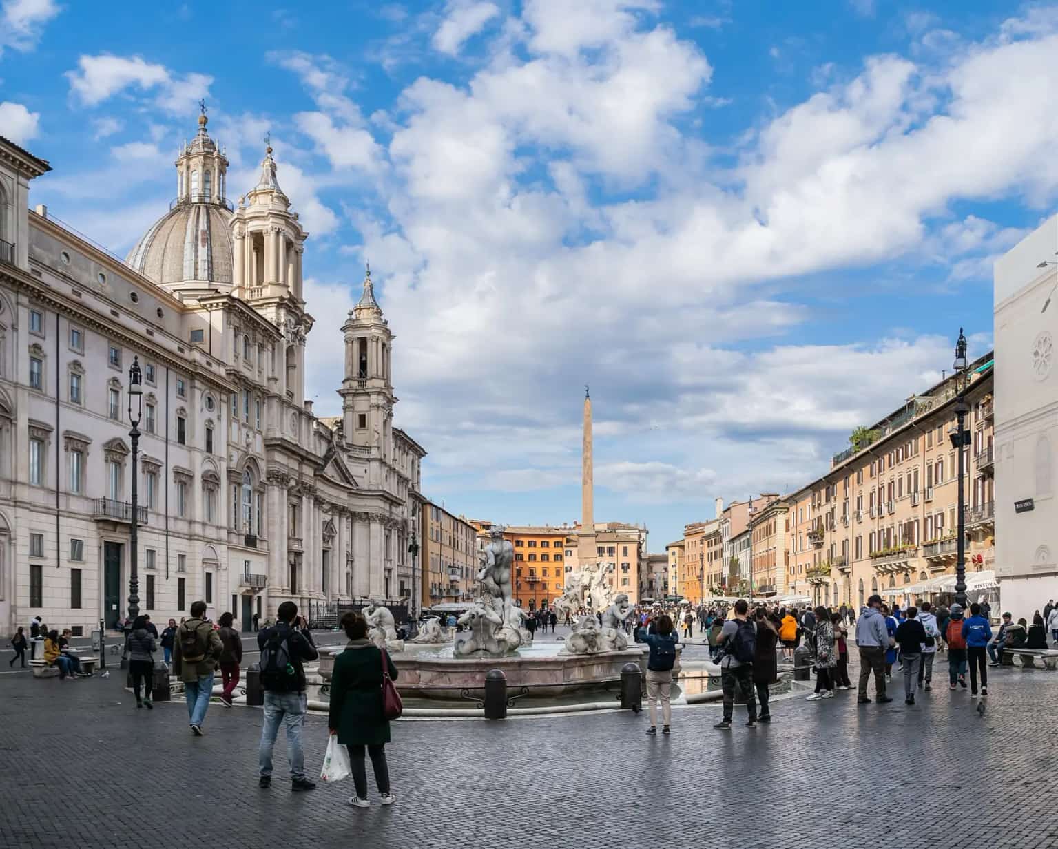 The long paving of Piazza Navona with the Four Rivers fountain and obelisk, the domed church of Sant'Agnese and strollers under a bright sky.