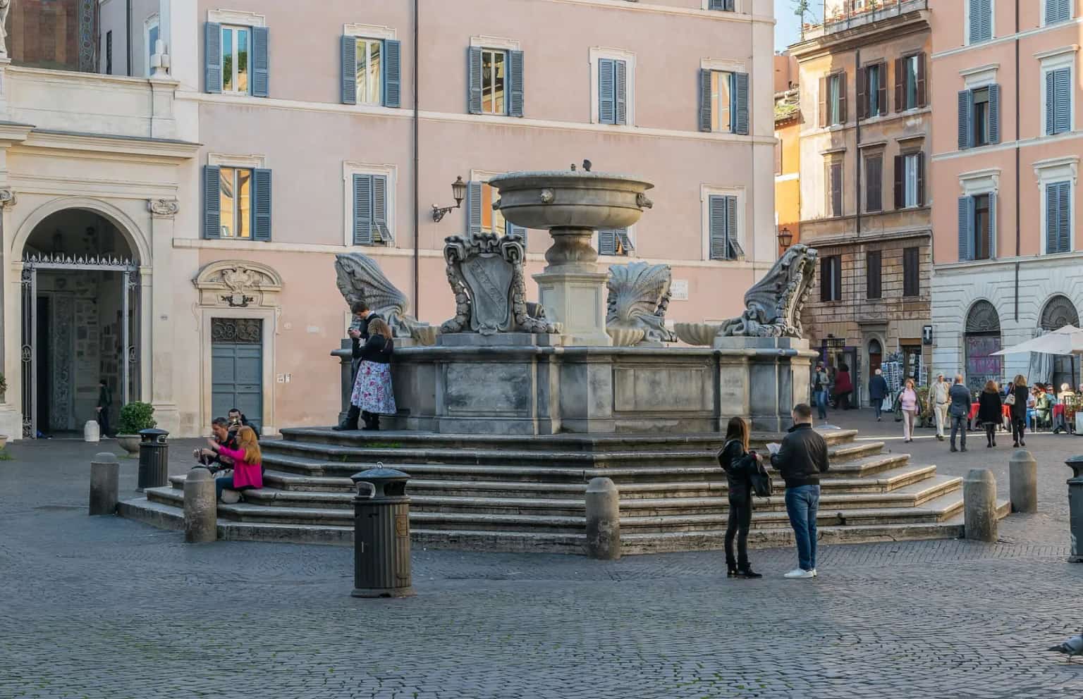 The stepped fountain in Piazza di Santa Maria in Trastevere with pastel buildings and people sitting on the steps.