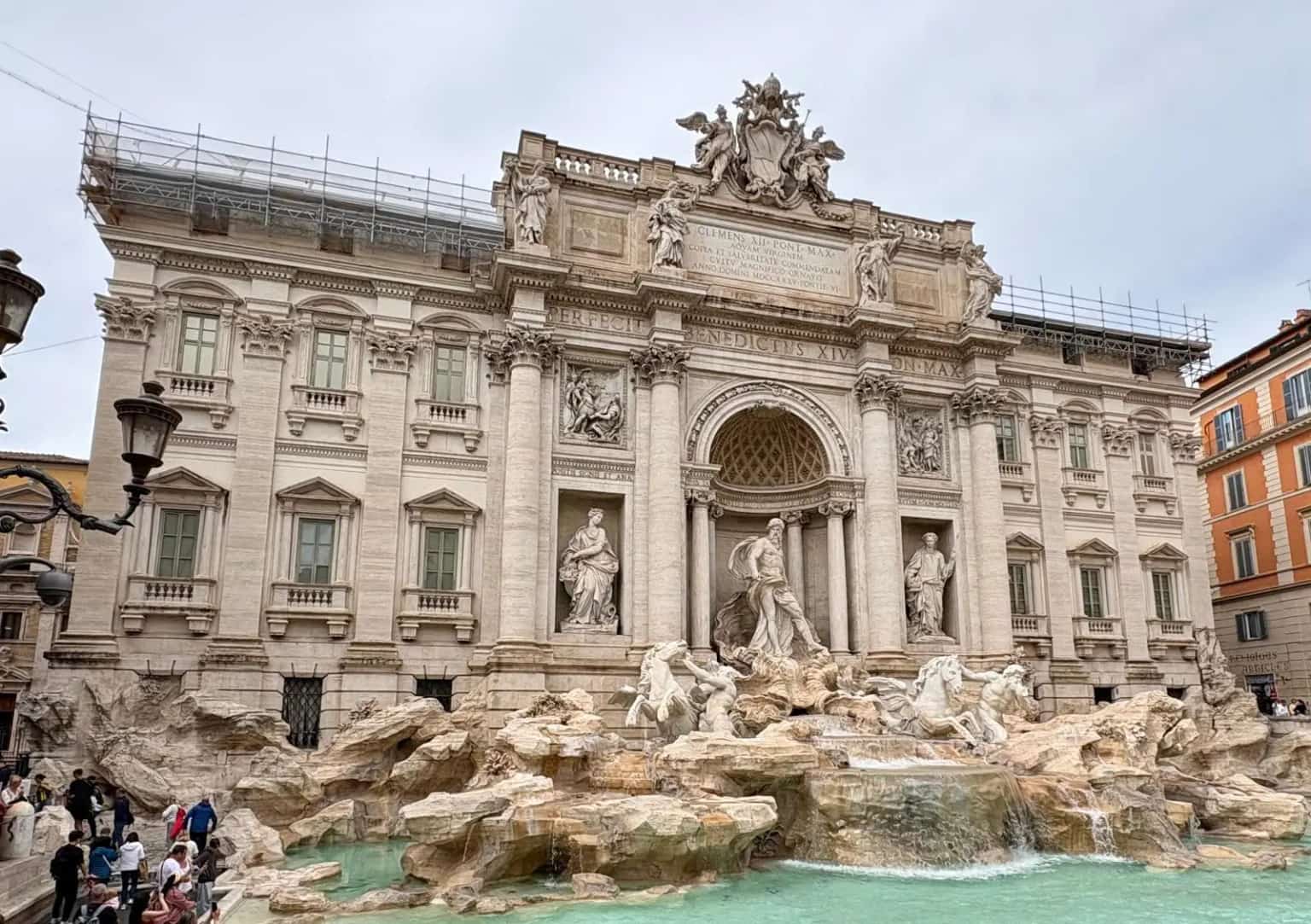 The Trevi Fountain's white marble facade and statues above its turquoise pool, with visitors at the edge.