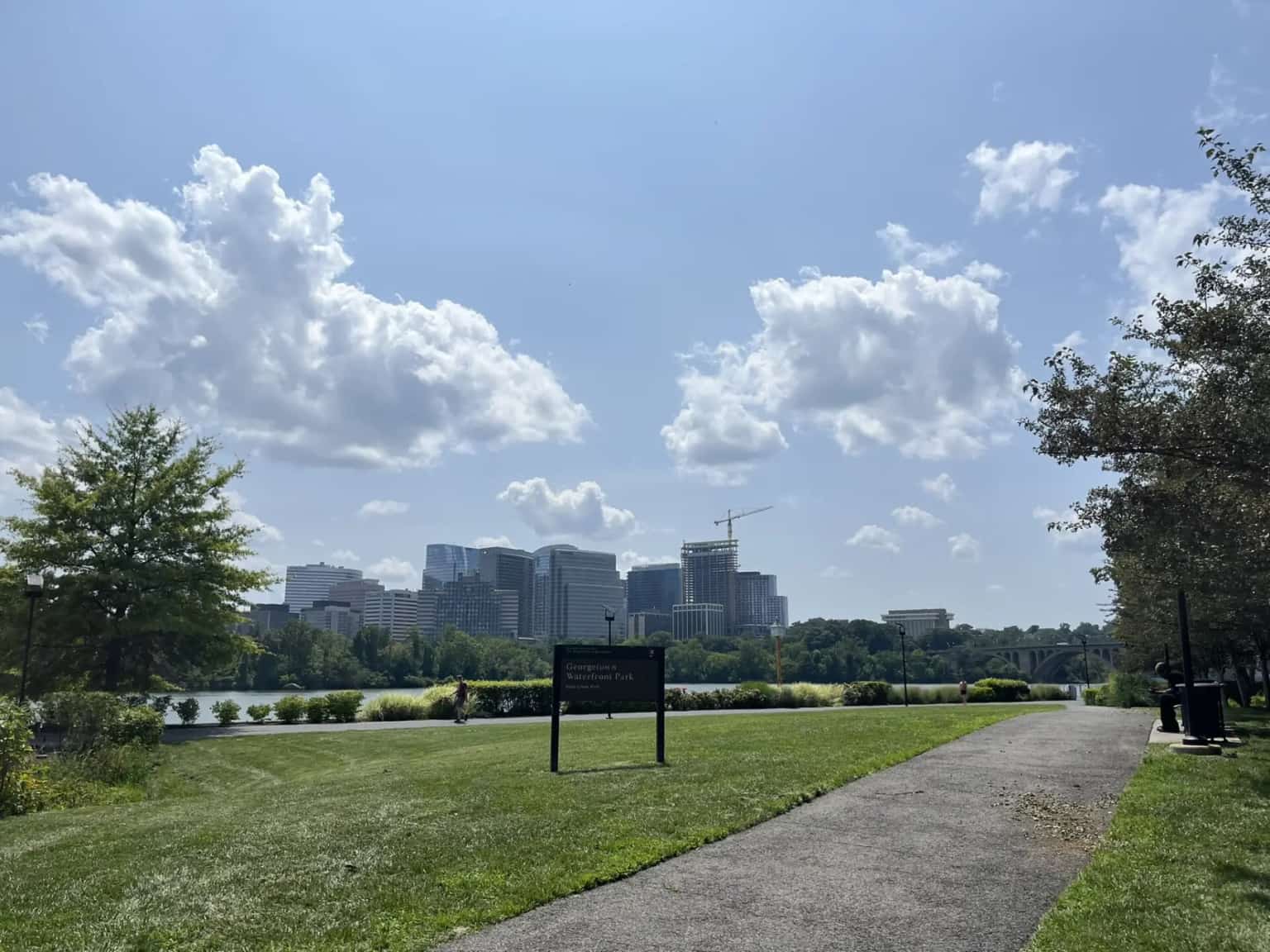 A paved path through green lawns at Georgetown Waterfront Park, the Potomac and Rosslyn towers beyond under white clouds.