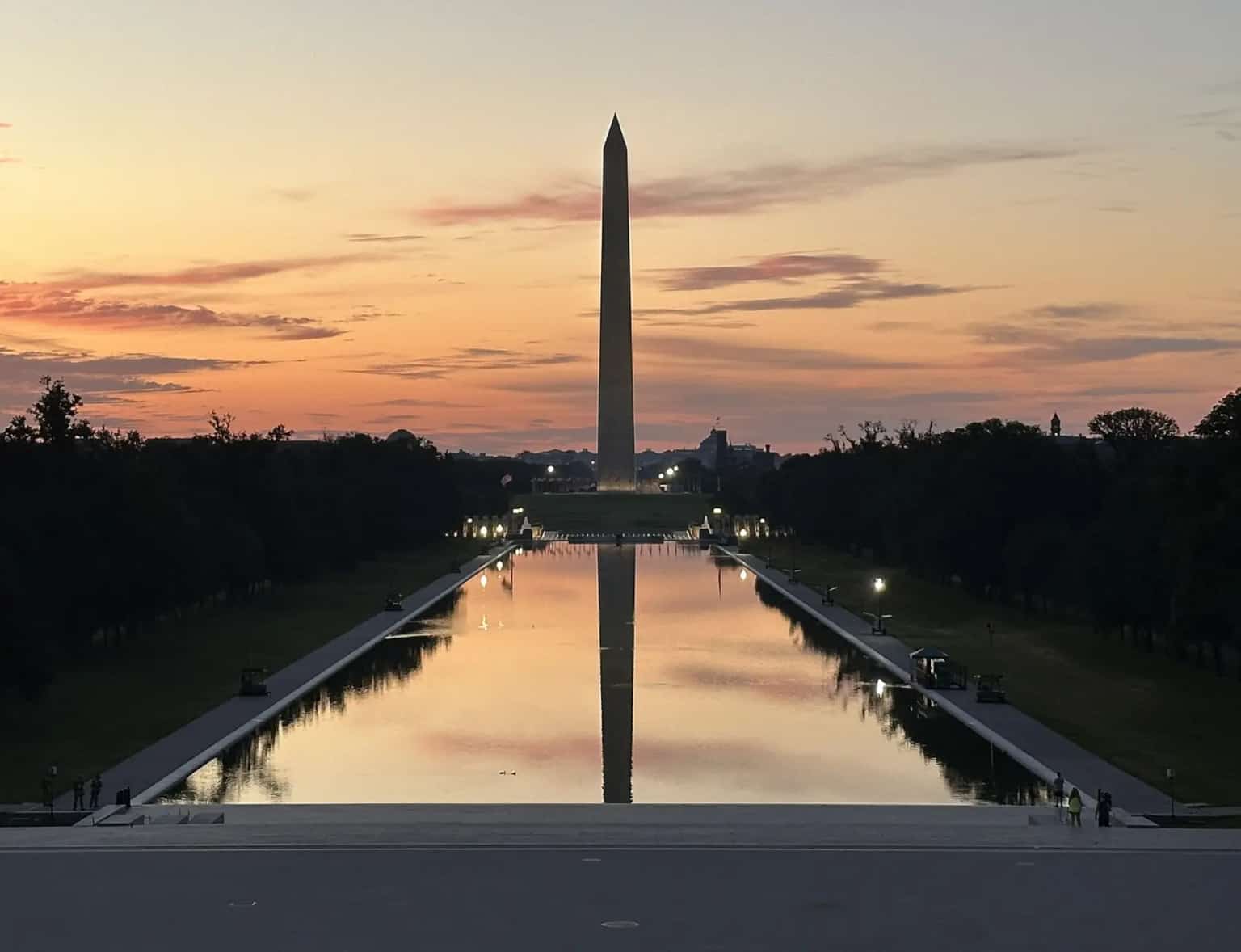 The Washington Monument and its reflection in the Reflecting Pool at sunrise, seen from the Lincoln Memorial steps under a pink sky.