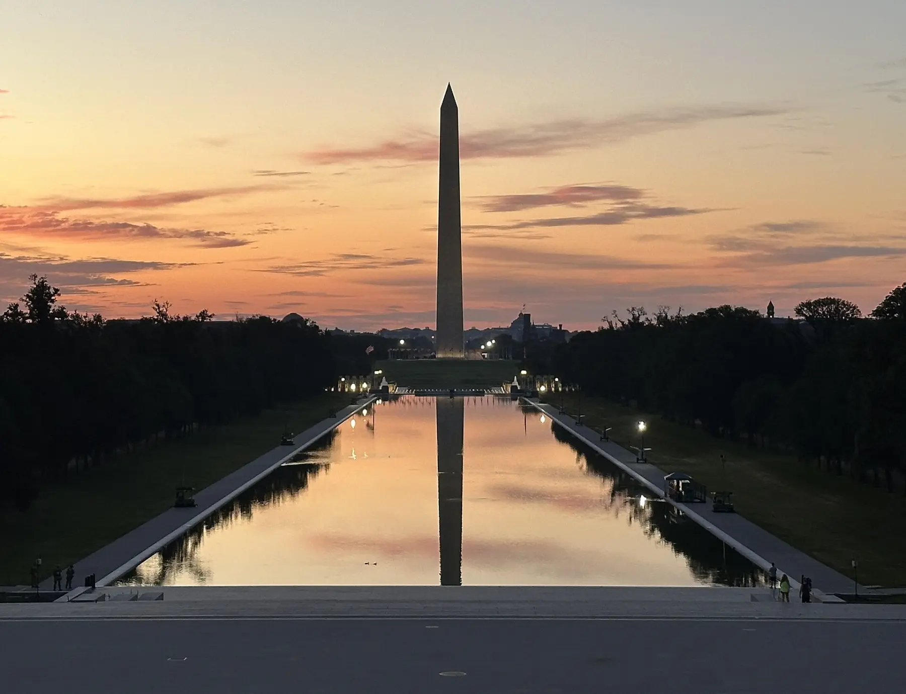 The Washington Monument and its reflection in the Reflecting Pool at sunrise, seen from the Lincoln Memorial steps under a pink sky.