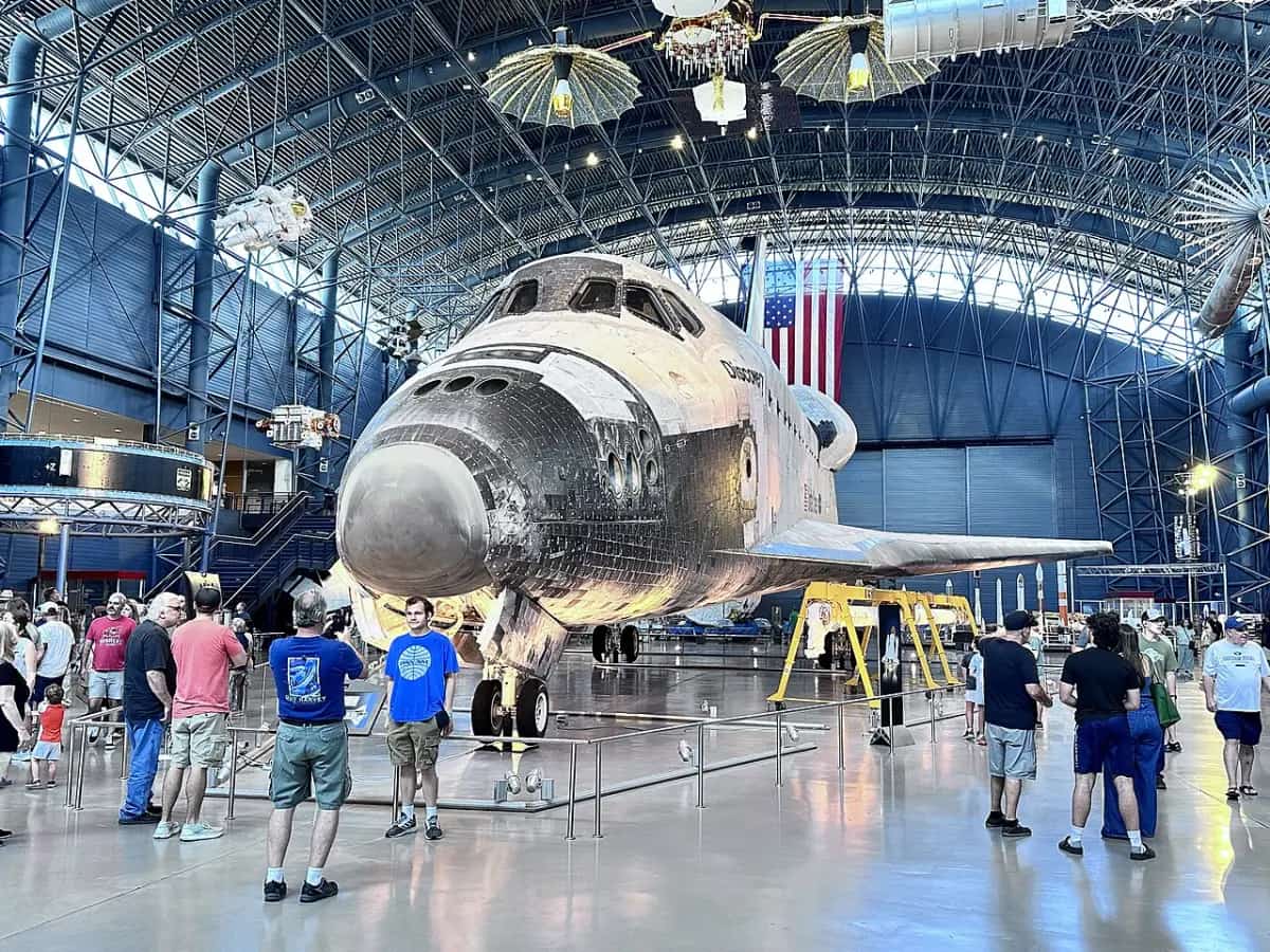 The Space Shuttle Discovery inside a vast hangar at the Udvar-Hazy Center, with visitors walking around it.