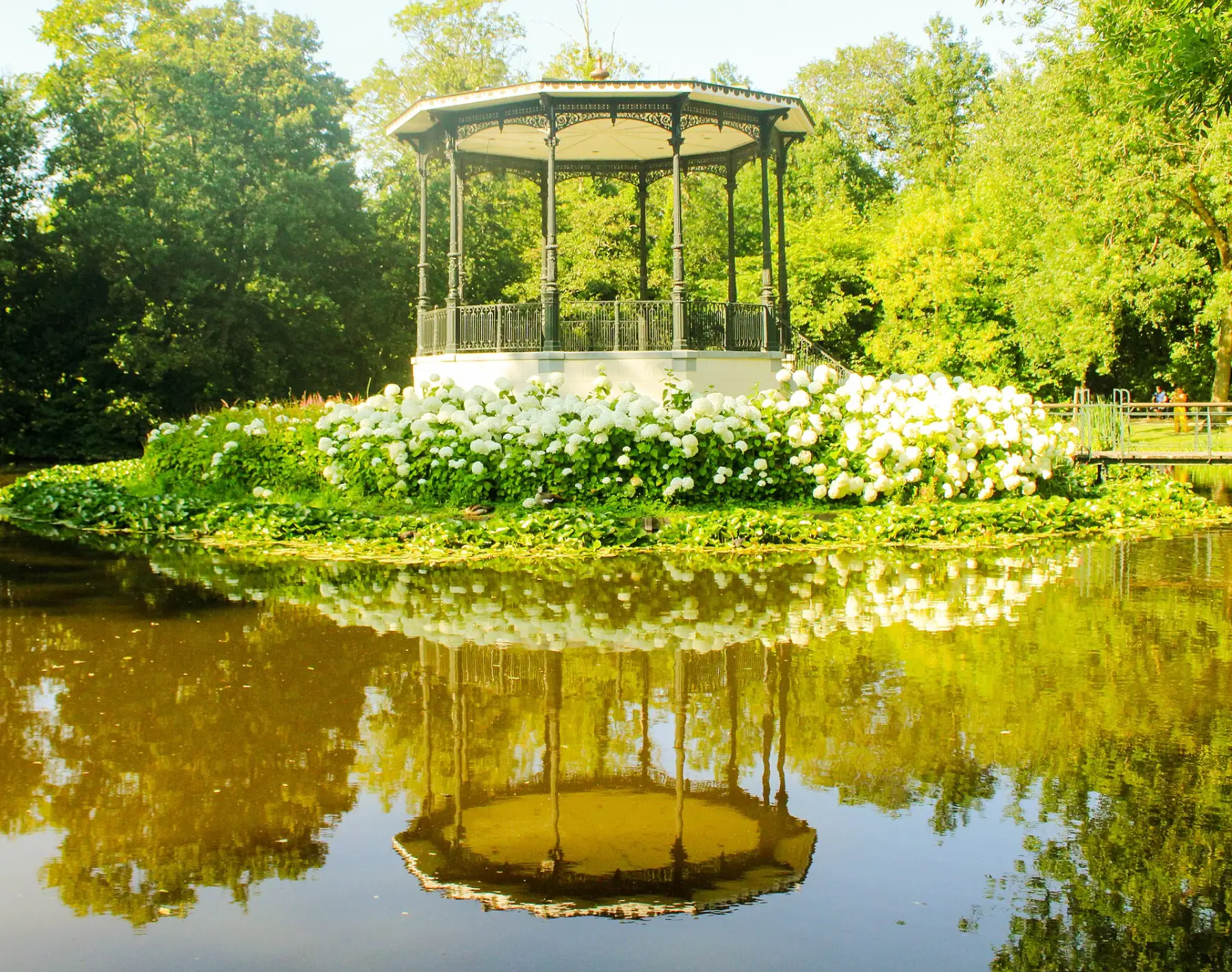 The white bandstand in the Vondelpark on a small island, reflected in a still pond surrounded by bright green trees.