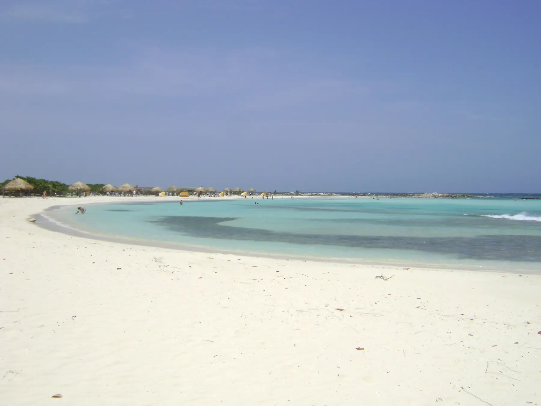 The curved white sand and pale turquoise shallow lagoon of Baby Beach with thatched shade huts in the distance