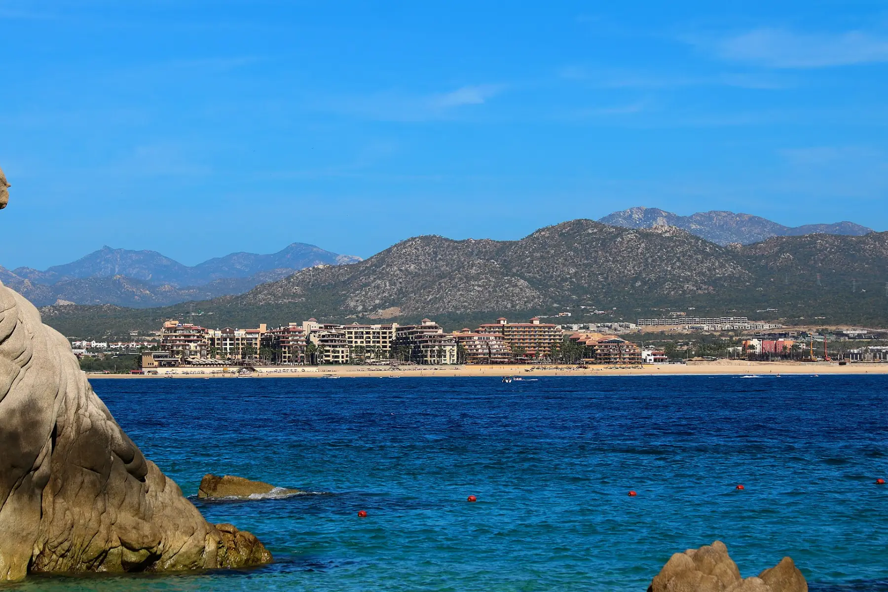 Medano Beach seen from the bay, a strip of hotels along the sand with desert mountains behind and deep blue water in front