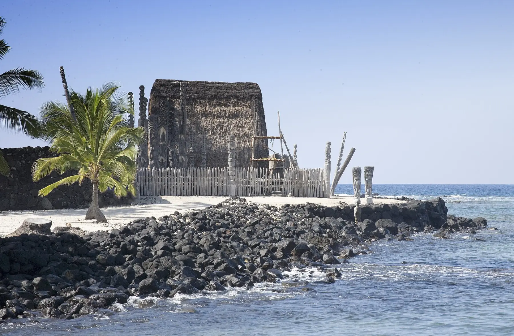 A thatched temple and carved wooden images on black lava rock beside the sea at Puʻuhonua o Hōnaunau