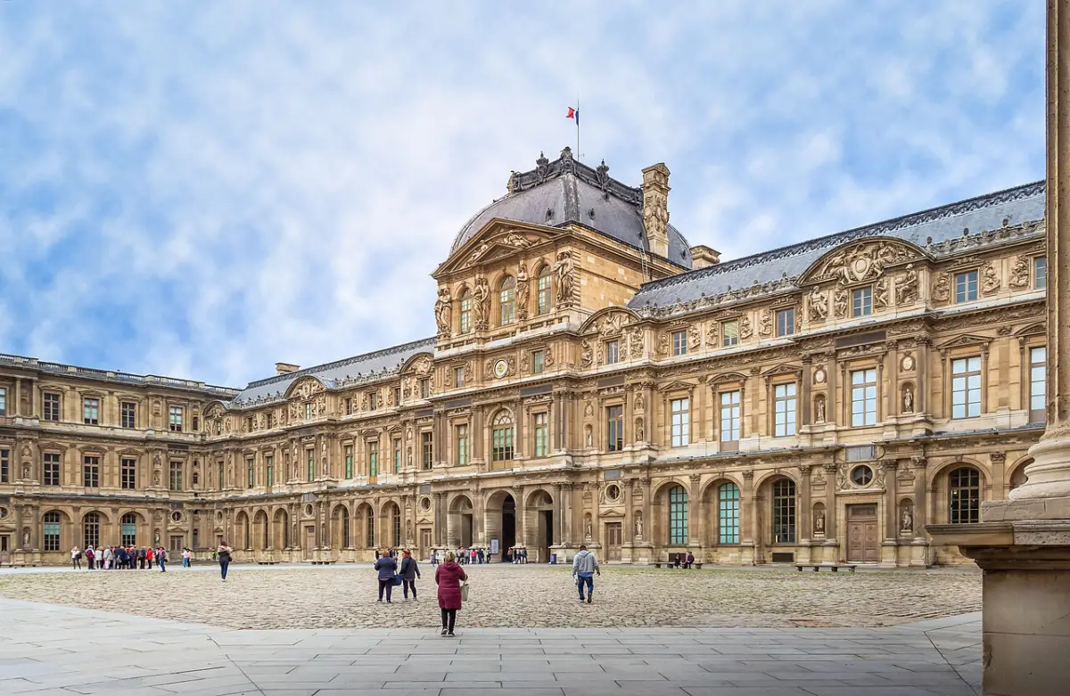 The ornate west facade of the Louvre's Cour Carrée courtyard under a bright sky, with a few visitors crossing the paving.